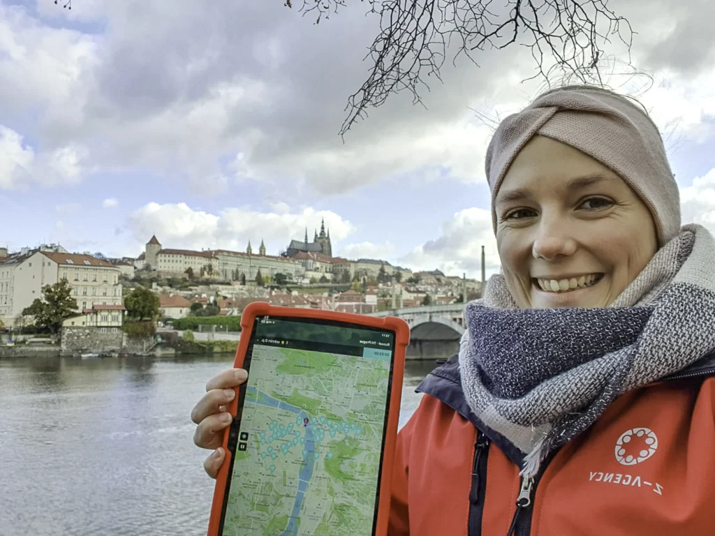 Prowadząca grę Treasure Hunt z zespołu Z-AGENCY z tabletem na nabrzeżu Wełtawy w Pradze, w tle Zamek Praski na drugim brzegu rzeki